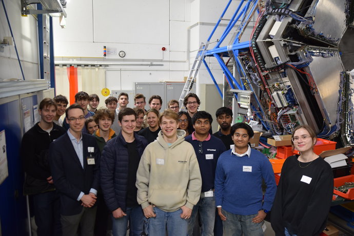 A group of persons stands in a large room. At the right-hand corner a huge experiment setup is visible.