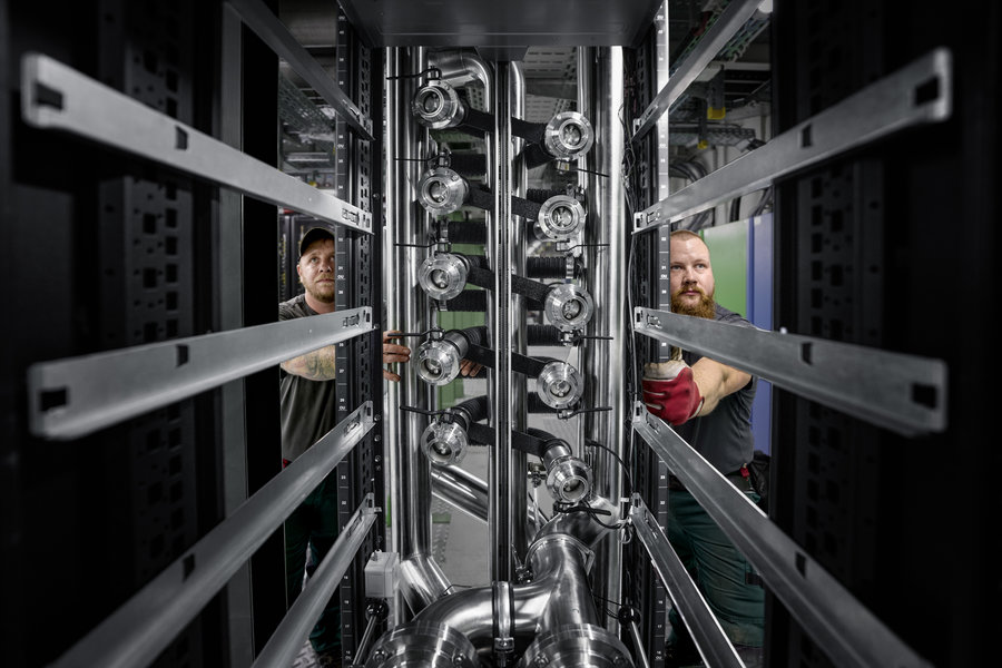 KI-System beim Einbau im Green IT Cube Perspektivisch durch ein schwarzes Computerrack geschaut, sind zwei Menschen sichtbar, die an Rohrleitungen arbeiten.