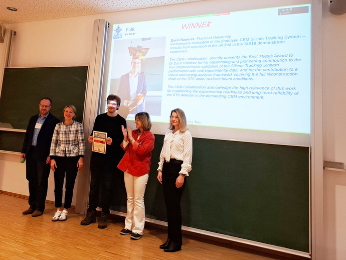 Five persons stand in a lecture hall in front of a blackboard. In the background, a presentation is visible.