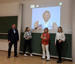 Vier Personen stehen in einem Vortragsraum vor einer Tafel. Im Hintergrund an der Wand ist eine Präsentation zu sehen.