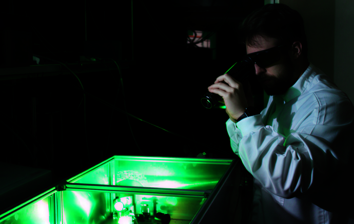 A man with protective laser-googles and white protective suit observes a green-glowing box through an optical measurement device.