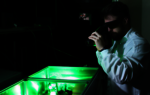Ein Mann mit Laserschutzbrille und weißer Schutzkleidung blickt durch ein optisches Messgerät auf eine grün leuchtende Box.