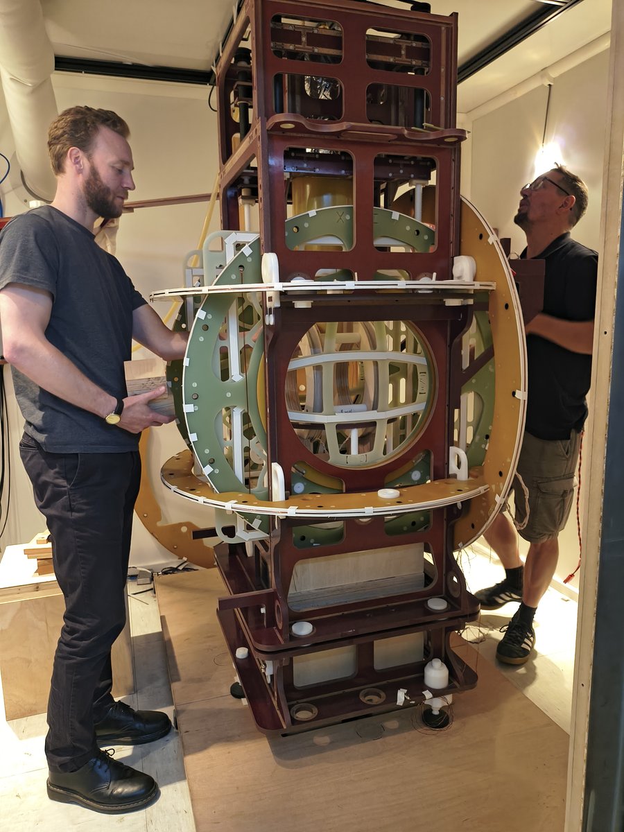 Zwei Männer stehen rechts und links von einem überkopfhohen, verschachtelten Holzgerüst.