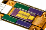 A detector module for the ECHo experiments developed and built at the Kirchhoff Institute for Physics. Auf einem kupferfarbenen Board ist mittig ein rechteckiger Mikrochip mit goldenen Verdrahtungen zu sehen. Er ist umgeben von vier kleineren grünen und blauen Chips.