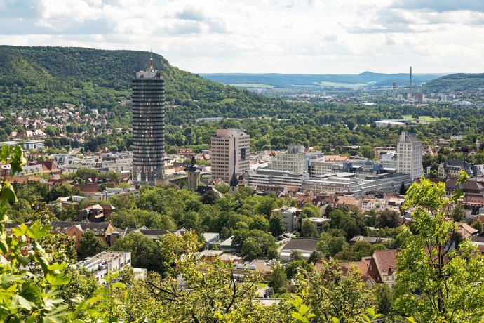 Von einem erhöhten Aussichtspunkt erkennt man die darunter liegende Stadt mit viel Grün, einem großen Turm und mehreren Gebäuden. Von einem erhöhten Aussichtspunkt erkennt man die darunter liegende Stadt mit viel Grün, einem großen Turm und mehreren Gebäuden.