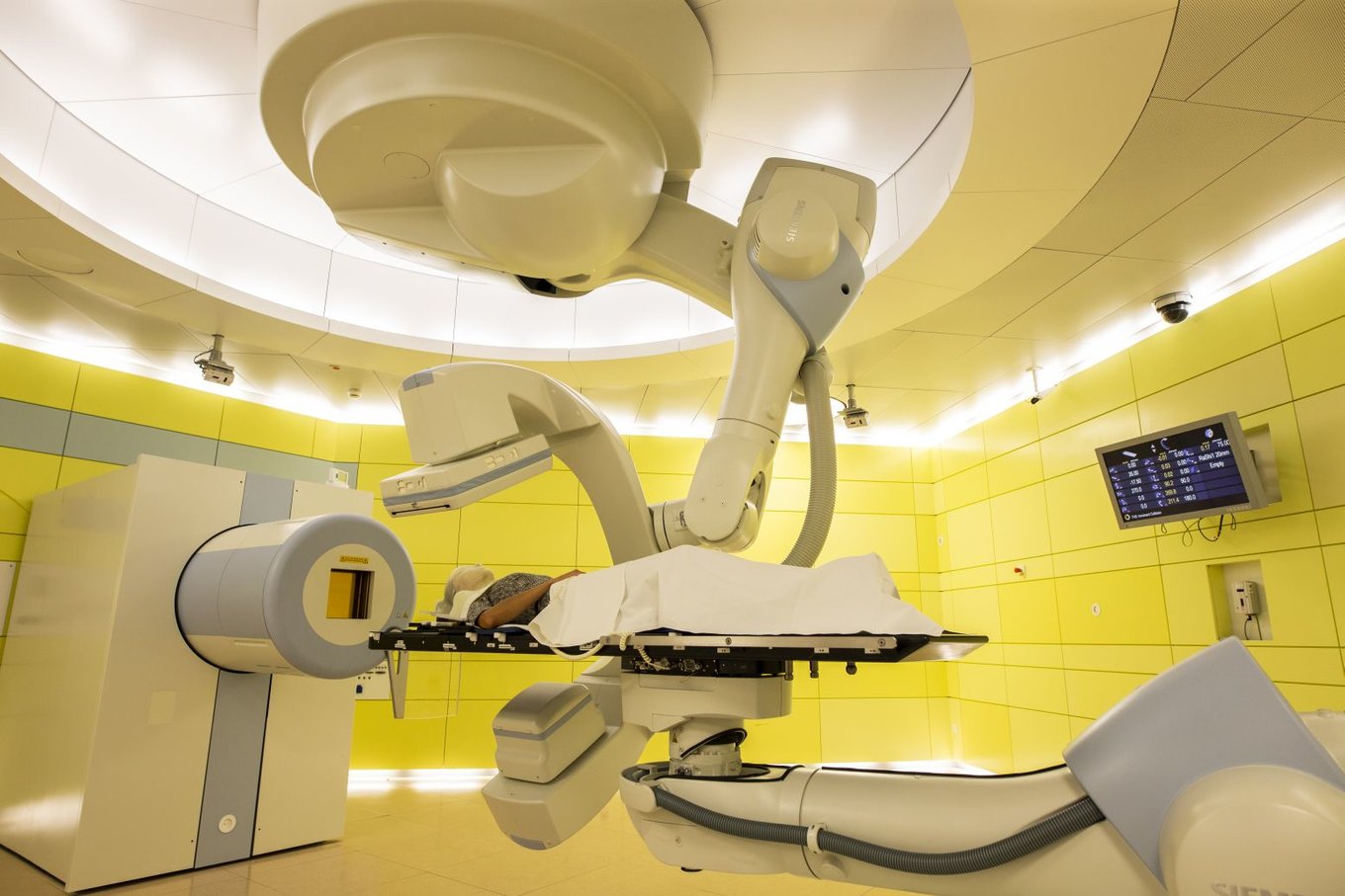 Eine Person liegt auf einer von einem Roboter gehaltenen Patientenliege vor einem zylindrischen Strahlaustrittsfenster.