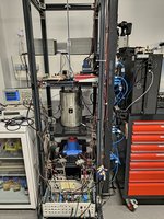 In einem Metallregal steht ein silberner Zylinder, darunter und darüber Messgeräte und Kabel.