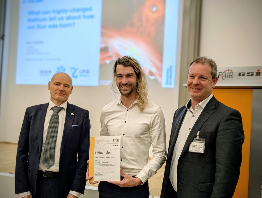 Preisübergabe des FAIR-GSI-Promotionspreises 2025 Drei Männer stehen vor einer Bühne, der mittlere hält eine Urkunde in der Hand.