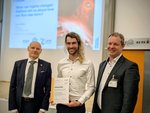 Preisübergabe des FAIR-GSI-Promotionspreises 2025 Drei Männer stehen vor einer Bühne, der mittlere hält eine Urkunde in der Hand.
