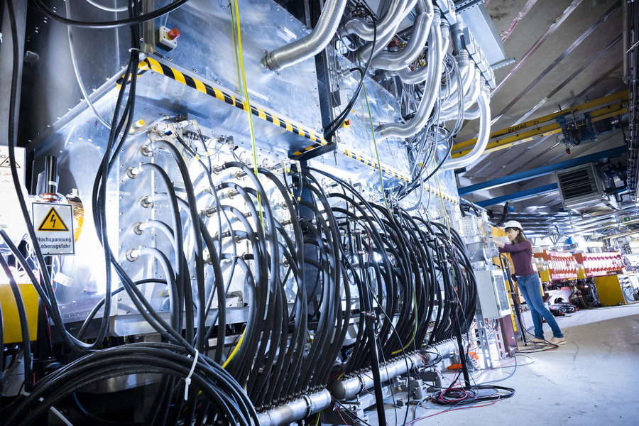 Beschleunigerkavität am Ringbeschleuniger SIS18 von GSI/FAIR Eine Frau steht neben einer silbernen Box, in die eine Vielzahl von schwarzen Kabeln hineinführt.